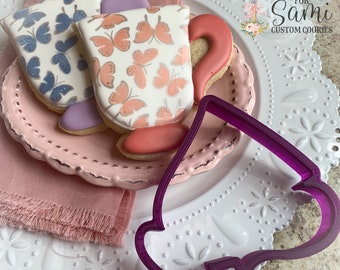 May include: Two decorated sugar cookies in the shape of teacups with butterfly designs. A purple plastic cookie cutter in the shape of a teacup is on a white plate with a pink lace border.