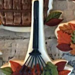May include: A white cookie decorated with a black rake and colorful fall leaves. The rake has a wooden handle and metal tines. The leaves are red, orange, yellow, and green.