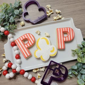 May include: A white rectangular tray holds cookies spelling "POP" and a popcorn-shaped cookie. The "P" cookies have red and white stripes. A purple popcorn-shaped cookie cutter and popcorn kernels are also present. A red and white bead garland is on the left.
