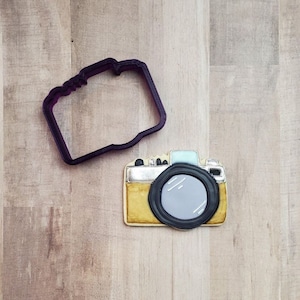 May include: A purple plastic cookie cutter shaped like a vintage camera, next to a decorated cookie shaped like a camera. The cookie is brown with a silver lens and a black lens.