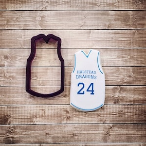 May include: A white cookie shaped like a basketball jersey with the text "Halstead Dragons" and the number "24" in blue. A purple plastic cookie cutter shaped like a basketball jersey is next to the cookie.