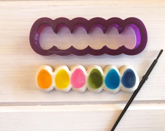 May include: A purple plastic cookie cutter shaped like six connected eggs, next to a row of six decorated cookies shaped like eggs, with a black paintbrush.