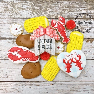 May include: A set of decorated sugar cookies featuring a red lobster, corn on the cob, a heart-shaped cookie with two lobsters, and a wooden sign that reads "Whitney & Alicia".