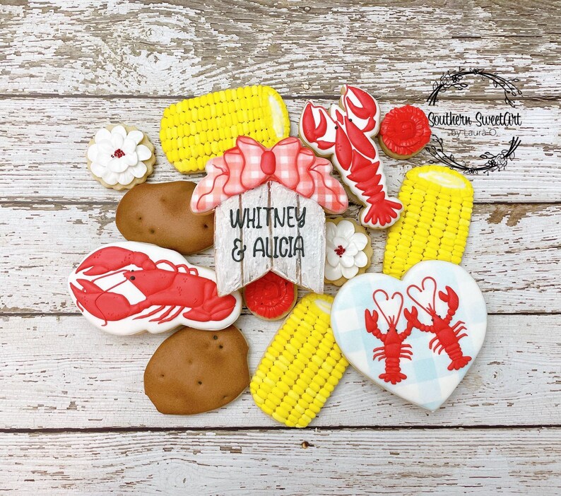 May include: A set of decorated sugar cookies featuring a red lobster, corn on the cob, a heart-shaped cookie with two lobsters, and a wooden sign that reads "Whitney & Alicia".