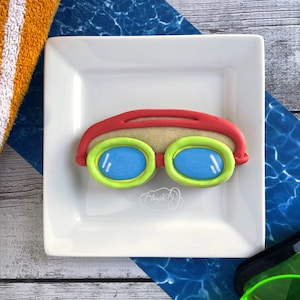 May include: A red, white, and blue decorated sugar cookie shaped like a pair of swimming goggles. The goggles have green trim and blue lenses.