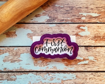 Op de afbeelding: Paarse plastic koekjesvormer in de vorm van een wolk met de woorden "First Communion" in een cursief schrift.