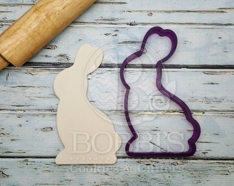 May include: A wooden rolling pin rests near a bunny-shaped cookie cutter and a bunny-shaped cookie. The cookie cutter is purple, and the cookie is off-white. The background is a light blue, weathered wood surface. The cookie cutter and cookie are for Easter.
