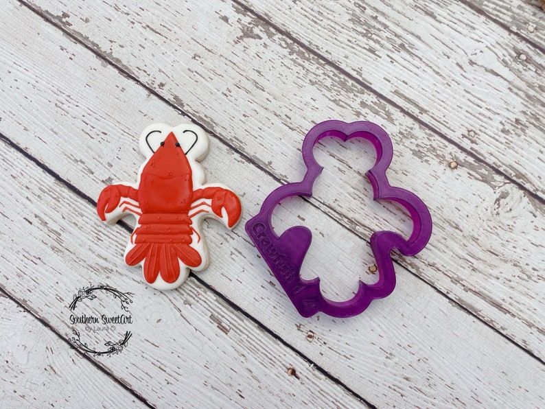 K&ouml;nnte beinhalten: Ein roter und wei&szlig;er Hummer-Keks mit einem violetten Plastik-Ausstecher in Form eines Hummers. Der Ausstecher ist mit "Crawfish" beschriftet.