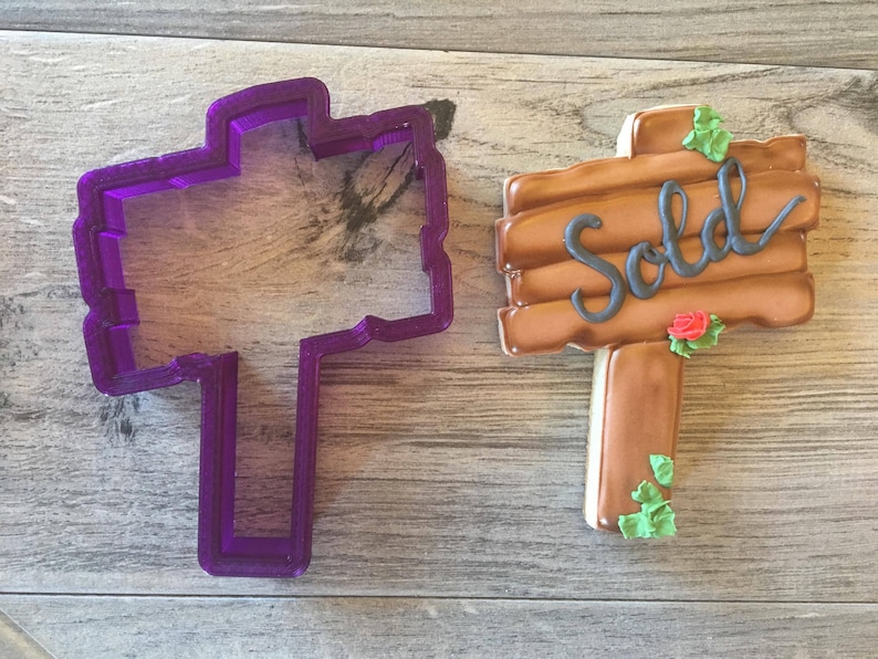 May include: Purple plastic cookie cutter shaped like a wooden sign with the word "Sold" on it. Next to the cutter is a decorated cookie in the shape of a wooden sign with the word "Sold" on it.