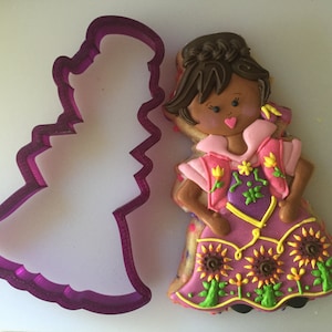 May include: Purple cookie cutter shaped like a dress, next to a decorated sugar cookie of a girl wearing a pink and purple dress with sunflowers on the bottom.