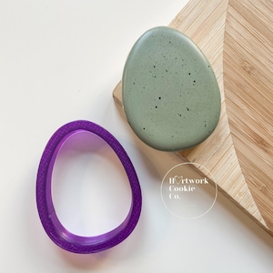 May include: A purple plastic cookie cutter shaped like an egg, next to a green speckled egg-shaped cookie on a wooden cutting board. The text "Heartwork Cookie Co." is visible on the cutting board.