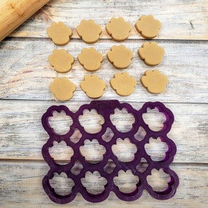 May include: A purple plastic paw-shaped cookie cutter with 12 individual paw prints.  There are 12 brown paw-shaped cookies on a wooden surface next to the cookie cutter.