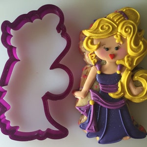 May include: Purple plastic cookie cutter shaped like a woman with long hair. The cutter is next to a decorated sugar cookie of a woman with long yellow hair, a purple dress, and a red heart on her face.