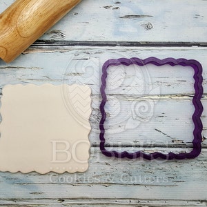 May include: A purple plastic cookie cutter in the shape of a square with a scalloped edge. The cutter is on a light blue wooden surface with a rolling pin.