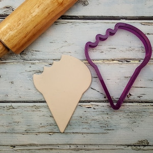 May include: A purple plastic ice cream cone cookie cutter with a bite taken out of it, next to a beige clay ice cream cone cookie.