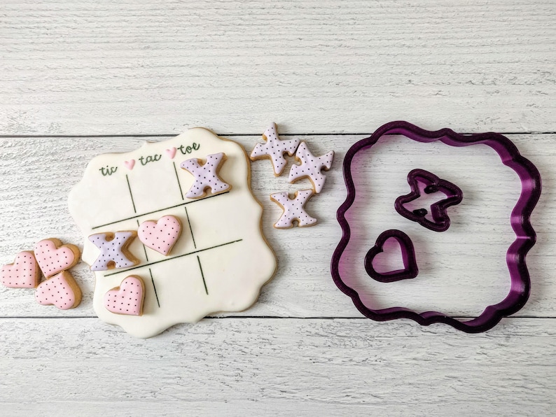 May include: A white tic-tac-toe board with pink and purple heart and X cookies. There are also purple cookie cutters in the shape of a heart and an X. The cookies and cutters are on a white wooden surface.