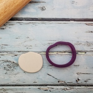 May include: A close-up of a beige circle of clay dough next to a purple plastic cookie cutter on a light blue wooden surface.