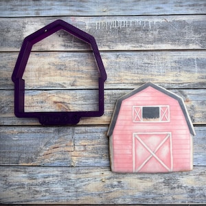 May include: A pink barn cookie with a white X on the door and a black window with hay sticking out. A purple plastic cookie cutter in the shape of a barn is next to the cookie.