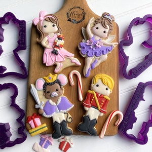 May include: Purple plastic cookie cutters shaped like a ballerina, a nutcracker, a mouse, and a candy cane. The cutters are on a wooden cutting board with decorated sugar cookies.