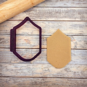 May include: Purple plastic cookie cutter shaped like a pointed shield. A brown cookie is cut out using the cookie cutter.
