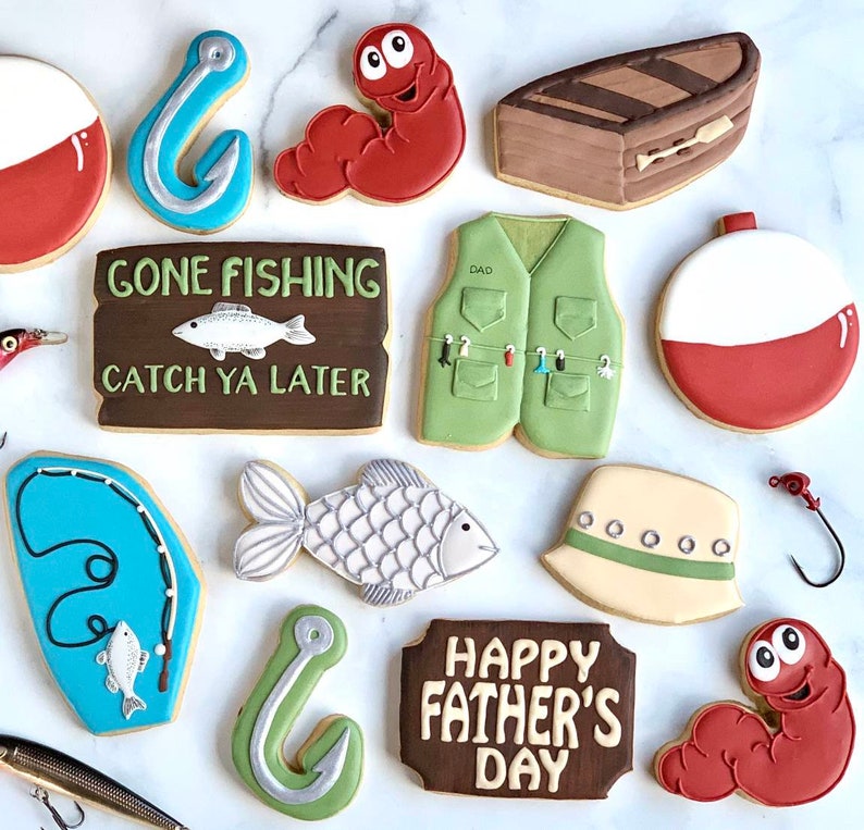 May include: A collection of decorated sugar cookies with a fishing theme. Cookies include a fishing vest, bobbers, fish, fishing hooks, a boat, and worms. Some cookies have text that reads "Gone Fishing Catch Ya Later" and "Happy Father's Day."