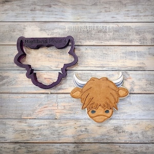May include: A purple plastic cookie cutter in the shape of a Highland cow head, with the text "Highland Head 4". Next to it is a decorated cookie in the shape of a Highland cow head, with brown fur, white horns, and blue eyes.