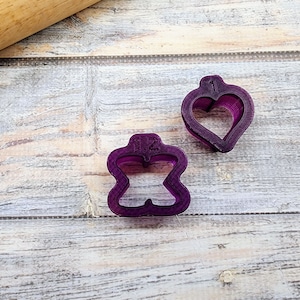 May include: Two purple plastic cookie cutters, one shaped like a heart with the number "1" on it, and the other shaped like a flower with the number "1/2" on it.