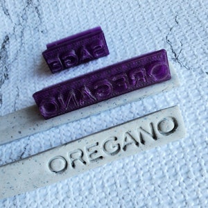 May include: Three purple rubber stamps with the words "Sage", "Oregano", and "Oregano" on a white speckled surface.