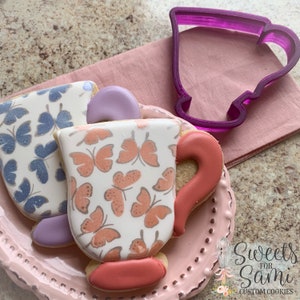 May include: Two decorated sugar cookies in the shape of teacups. The cookies are decorated with a butterfly pattern and have a purple and red handle. A purple plastic teacup cookie cutter is in the background.