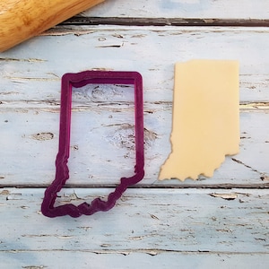 May include: A purple cookie cutter in the shape of the state of Indiana, next to a cut-out cookie in the same shape. A wooden rolling pin rests in the background on a distressed, light blue wooden surface.