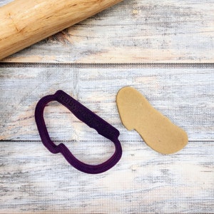 May include: Purple plastic cookie cutter shaped like a vintage car with the text "Ruby's Rolling Diner" on the side. A piece of cookie dough is cut out using the cookie cutter.