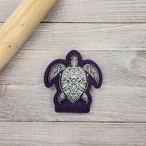 May include: Purple plastic cookie cutter shaped like a sea turtle with a geometric pattern. The cutter has the words "Sea Turtle" written on the bottom.