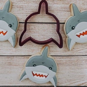 May include: Three gray and white shark-shaped cookies with orange teeth and black eyes. A purple shark-shaped cookie cutter is in the center.