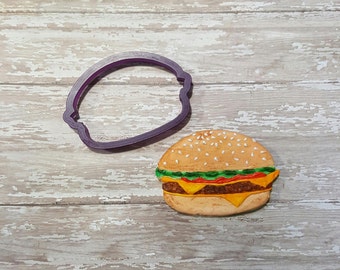 May include: A purple plastic cookie cutter shaped like a hamburger bun, next to a decorated sugar cookie shaped like a hamburger with sesame seeds, lettuce, tomato, cheese, and a meat patty.