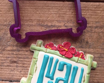 Puede incluir: Cortador de galletas de plástico morado en forma de marco rectangular con un borde superior festoneado. Una galleta decorada con un marco de bambú y la palabra "LUA" en glaseado azul se muestra junto al cortador.