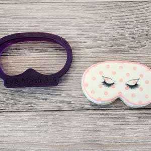 May include: Purple plastic cookie cutter shaped like a sleep mask with the text "Eye Mask 3 S" on the bottom. Next to the cutter is a pink and white decorated cookie shaped like a sleep mask with eyelashes and pink polka dots.