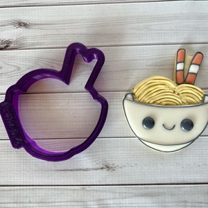 May include: Purple plastic cookie cutter shaped like a bowl of ramen noodles with chopsticks.  Next to the cutter is a decorated cookie of a bowl of ramen noodles with chopsticks.