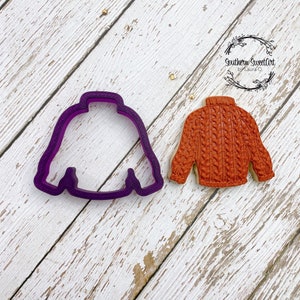 May include: A purple plastic cookie cutter and a brown sugar cookie in the shape of a sweater.  The cookie cutter is on the left and the cookie is on the right.  The cookie is decorated with a cable knit pattern.