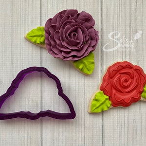 May include: Two sugar cookies decorated with purple and red frosting in the shape of roses with green leaves. A purple plastic cookie cutter in the shape of a rose is also shown.