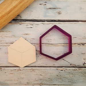 May include: A close-up of two cookie cutters on a white wooden surface. The cookie cutters are made of plastic and are shaped like a hexagon and a cube. The hexagon cookie cutter is purple and the cube cookie cutter is beige.
