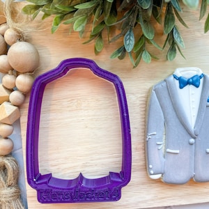 May include: Purple plastic cookie cutter shaped like a suit jacket with the words "Mens Jacket" on the bottom. A gray cookie decorated to look like a suit jacket is shown next to the cutter.