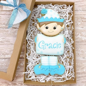 May include: A blue and white elf cookie with a sign that says "Gracie" in a brown cardboard gift box with white shredded paper.