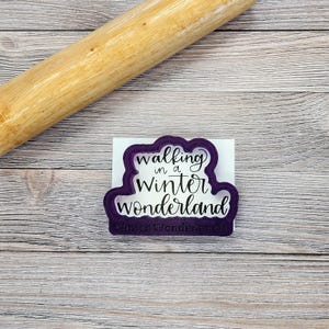 May include: A purple cookie cutter with the words "walking in a Winter Wonderland" in a script font. The cutter is on a white surface, with a wooden rolling pin in the background.