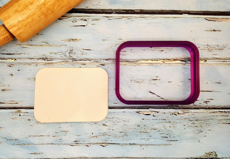 Puede incluir: Primer plano de dos cortadores de galletas sobre una superficie de madera. Los cortadores de galletas son ambos de forma cuadrada. Uno es de color marr&oacute;n claro y el otro es de color morado brillante.