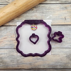 May include: A set of three purple plastic cookie cutters. The largest cutter is a square with rounded corners and a heart-shaped cutout. The medium cutter is a heart shape. The smallest cutter is a butterfly shape.