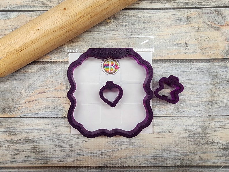 May include: A set of three purple plastic cookie cutters. The largest cutter is a square with rounded corners and a heart-shaped cutout. The medium cutter is a heart shape. The smallest cutter is a butterfly shape.