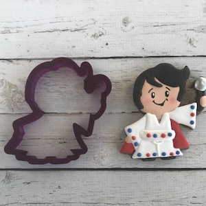May include: A purple plastic cookie cutter shaped like a person wearing a cape and holding a microphone. Next to the cutter is a decorated sugar cookie of a person wearing a white jumpsuit with red and blue accents, holding a microphone.