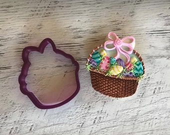 May include: A purple plastic cookie cutter next to a decorated Easter basket biscuit. The basket is brown with colourful eggs, green grass, and a pink bow. The eggs are decorated with various patterns and colours.