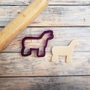 May include: Purple plastic cookie cutter shaped like a pig, next to a beige cookie in the shape of a pig, on a wooden surface with a wooden rolling pin.