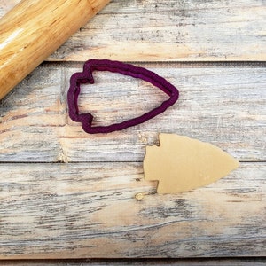 May include: A purple plastic arrowhead cookie cutter and a brown arrowhead cookie on a wooden surface. A wooden rolling pin is in the background.